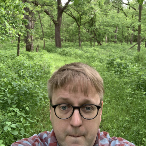 Photo of Nolan Tredway, facing the camera in front of a back drop of green forest